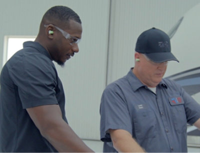 Dva muži v pracovních uniformách a sluchátkách diskutují o něčem v leteckém prostředí.