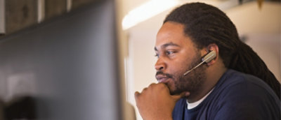 Çenesine mikrofon takılı bir kulaklık takmış, bilgisayar ekranına bakan bir erkek.