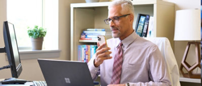 En mann i hvit skjorte som holder en telefon og ser på en bærbar datamaskin.