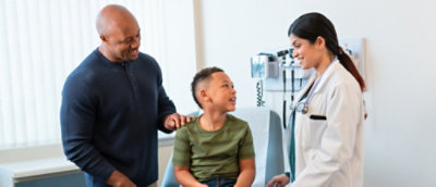 Un bărbat în cămașă albastră și o femeie în haină albă zâmbesc la un copil îmbrăcat în cămașă verde.