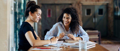Two women sitting at a table with one writing on a paper.