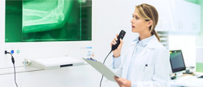 Un médecin en blouse blanche tenant un bloc-notes et un dispositif de dictée, examinant une radiographie du bras affichée sur un moniteur dans un cabinet médical.