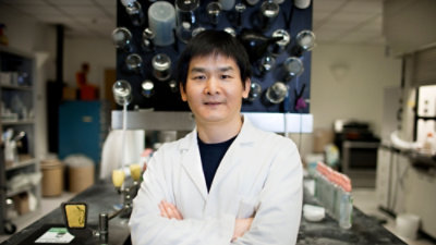 A person wearing a white lab coat in front of a plain background.