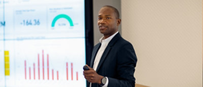 A person in a suit holding a microphone, standing in front of a screen displaying -164 36.