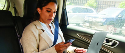 A woman sitting inside a car using smartphone and holding a laptop with other hand