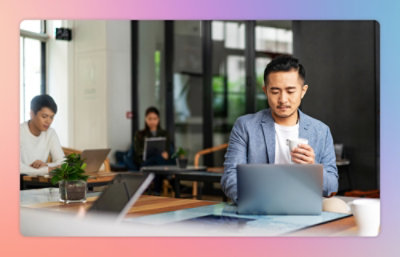 A man in a blue blazer uses a laptop while holding a cup in a modern office with several other people 