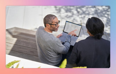 Two people sitting outdoors, one pointing at a laptop screen. The background is blurred with soft pastel hues.