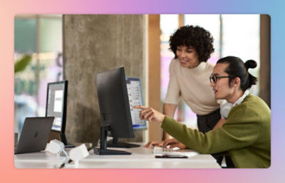 Two people are working together at a desk with desktop computers. One is seated, pointing at the screen, while the other stands