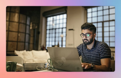 A man wearing glasses and a striped shirt works on a laptop at a desk in a modern room with large windows.