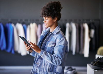 A person wearing a denim jacket uses a tablet in a clothing store, with racks of hanging clothes visible in the background.