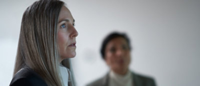 A woman with long straight hair and a serious expression faces forward, with another person blurred in the background.A woman with long straight hair and a serious expression faces forward, with another person blurred in the background.