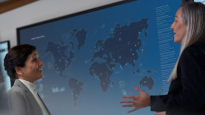 Two women in business attire converse in front of a large digital world map display filled with data points and charts.