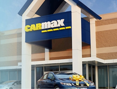A blue car with a yellow bow on it is parked in front of a building with a CARmax sign.