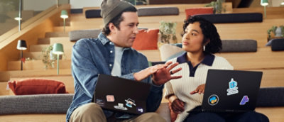 A man and woman are seated and engaged in a discussion while looking at laptops.