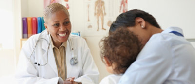A smiling doctor with a stethoscope talks to a parent holding a child in a medical office.