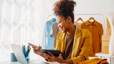A woman sits at a desk with a laptop and tablet, pointing at the screen.