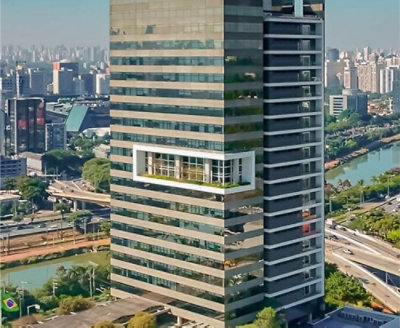 一棟高聳的現代玻璃辦公大樓，帶有外突的白色結構，周圍綠樹成蔭，遠處可見河畔城市天際線。