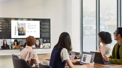 People are at a conference room table participating in a Teams call that is displayed on a large tv screen on the wall.