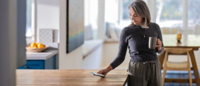 A person holding a phone and a coffee cup near a chair with a cushion, with a blurry window in the background.