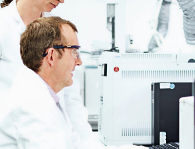 A man in a white lab coat and goggles looking at a computer.
