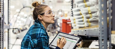 A person wearing safety goggles and a blue plaid shirt holding a tablet.