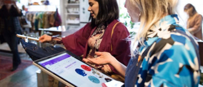 Two women are browsing a tablet in a clothing store, likely comparing clothing styles or making a purchase.