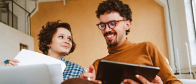 A man and woman sit on a couch, focused on a tablet they are sharing between them.