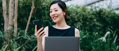 A woman smiles while holding a laptop in one hand and looking at her phone with the other.