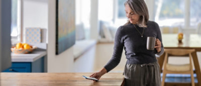 A woman stands by a kitchen table, holding a cup and looking towards the camera with a warm smile.