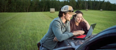 Two people sitting on a car hood, using a laptop, enjoying a casual outdoor work or leisure moment.