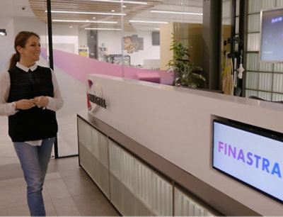 A person in a black vest walking past a desk with a screen displaying text and a potted plant nearby.