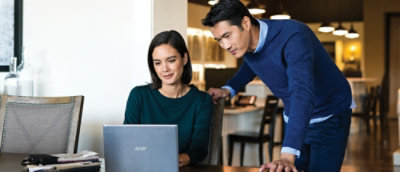 A man and a woman are standing next to each other looking at a laptop in a room with a black chair.
