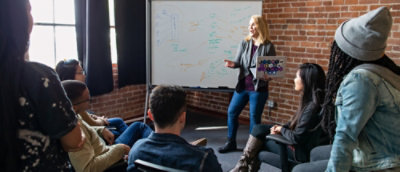 Group of people seated in a brick-walled room, attentively listening to a presenter pointing at a whiteboard
