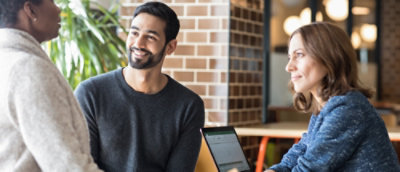 Three people indoors having a discussion, one using a laptop.