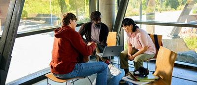 Three people sit in a bright room with large windows, engaging in discussion around a laptop.