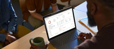 A person with a green mug uses a laptop displaying data dashboards, while two others sit nearby collaborating.