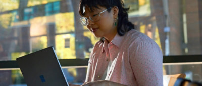 A person with glasses, wearing a pink shirt, works on a laptop in a sunlit room with large windows.