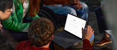 A diverse group of people sit in a circle, focused on a person using a laptop.