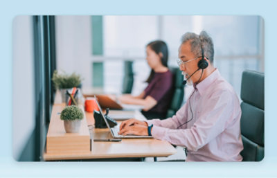 An man wearing a headset works on a laptop at a desk, with a woman working in the background in a modern office setting.