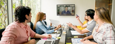 Group meeting around a table with video call on screen.