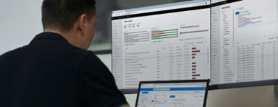 Person at desk with three monitors showing threat analytics, account logins, and messaging app.
