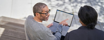 Two individuals sitting outdoors, one pointing at a laptop screen displaying text or a document.