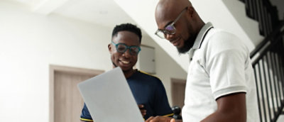 Two people standing indoors, one with a laptop and the other with a smartphone.