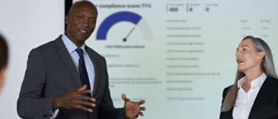 Two people in business attire talking in front of a screen with a gauge chart.