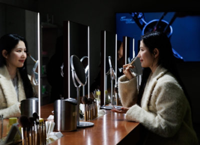 Žena sedí před zrcadlem a nanáší si štětcem make-up na obličej.