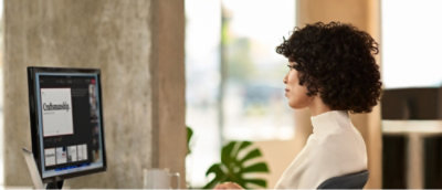 A woman sits at a desk, focused on her computer screen, with a notepad and pen beside her.
