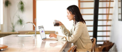 A women sitting at a table holding a cup of black cooffe looking at a laptop.