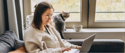 A women sitting on a couch working in a laptop and a cat behind her.