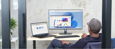 Person sitting at a desk with a large monitor displaying charts and graphs, and a laptop showing data,