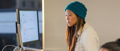 A close-up of a woman wearing a green beanie near a computer screen.